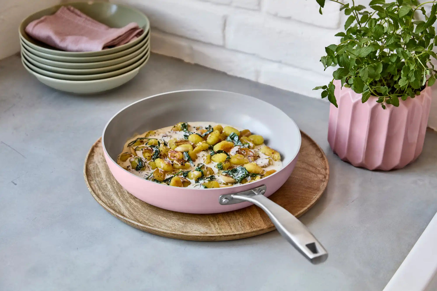 Růžová pánvička s lehce šedým vnitřkem a kovovou rukojetí, obsahující gnocchi a listovou zeleninu v krémové omáčce.